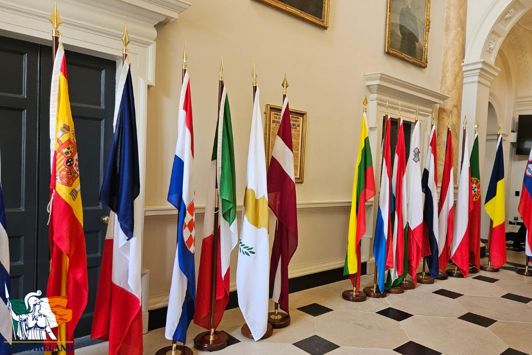 A row of international flags displayed on poles in an elegant indoor hall with cream walls, framed artwork, and a black-and-white checkered floor.