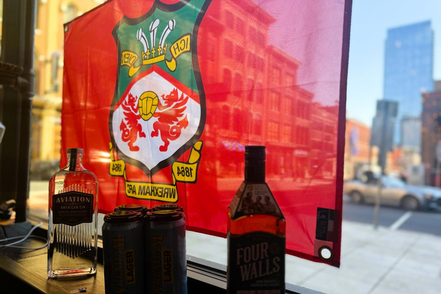 A red Wrexham AFC sports flag hangs on a window behind several bottles and cans of alcohol, including a bottle of Aviation and a bottle of Four Walls Lager.