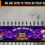 A Halloween-themed fence banner displayed on a black metal fence, featuring a purple spooky sky with bats, ghosts, pumpkins, and the text 'WE ARE OPEN TO TRICK OR TREAT BANNER'.