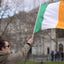 A woman in a brown jacket holds up a small Irish flag with green, white, and orange vertical stripes, with a blurred stone building and bare trees in the background.